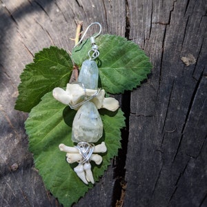 Op de afbeelding: Een unieke handgemaakte oorbel met een lichtblauwe kristal, kleine botjes en tanden, allemaal verpakt in zilverdraad. De oorbel hangt aan een zilveren haak. Gepresenteerd op een groen blad tegen een rustieke houten achtergrond.