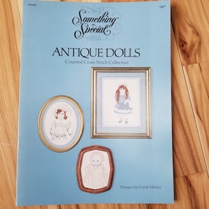 May include: A blue and white counted cross stitch pattern booklet featuring antique dolls. The cover shows three framed cross stitch designs of dolls in vintage clothing. The title is "Antique Dolls Counted Cross Stitch Collection" and the text "Designs by Candi Martin" is at the bottom.