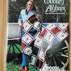 May include: A vintage book cover titled "Country Afghans" showing a woman with a patchwork afghan. The afghan has red, white, and blue squares with floral designs. The cover also displays the text "Cozy covers to knit and crochet."