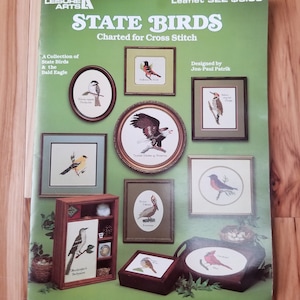 Puede incluir: Un folleto de patrones de punto de cruz con una colección de aves estatales y el águila calva. El folleto se titula "State Birds" e incluye el texto "Charted for Cross Stitch".