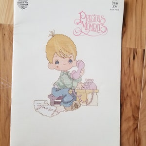 May include: Cross stitch pattern featuring a young boy with blonde hair sitting on a stool and holding a pink phone. The pattern is titled "Dear Jon" and is from the Precious Moments book PM-3.