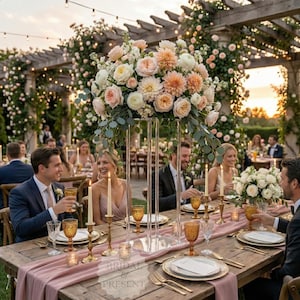 May include: A beautifully decorated outdoor wedding reception table with a pink runner, gold-rimmed plates, and amber glassware. A large floral arrangement with peach and cream-colored flowers sits atop a clear acrylic stand. Guests are seated around the table, enjoying the celebration.