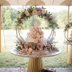 May include: A tiered wedding cake with a checkerboard pattern and floral decorations. The cake is surrounded by a gold hoop adorned with flowers and greenery. Cookies and other desserts are arranged on a marble-topped table.