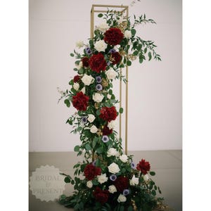 May include: A gold metal arch decorated with red peonies, white roses, and greenery. The arrangement includes small purple flowers and is accented with gold foliage. The words "Bridal & Present" are in the lower left corner.