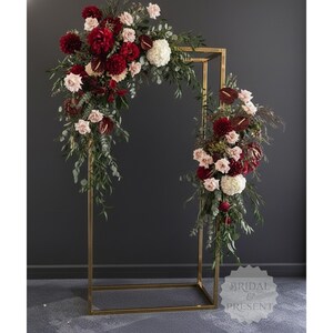 May include: A gold metal wedding arch decorated with an arrangement of red, white, and pink flowers and greenery. The arch is rectangular with a curved top, and the floral arrangement cascades down the sides. The text "BRIDAL & PRESENT" is visible.