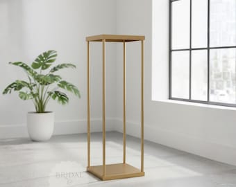 May include: A gold metal decorative stand with a square top and base, supported by four vertical bars. The stand is in a room with a white wall, a large window, and a potted plant. The stand is approximately 30 inches tall.