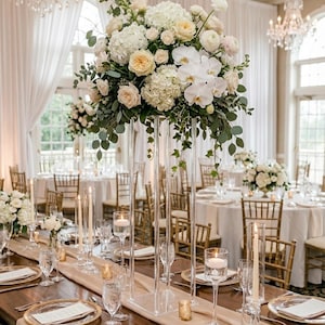 May include: A beautifully decorated wedding reception table with a tall, clear acrylic floral centerpiece. The arrangement features white and cream-colored flowers and greenery. The table is set with gold-rimmed plates, candles, and glassware, creating an elegant atmosphere.