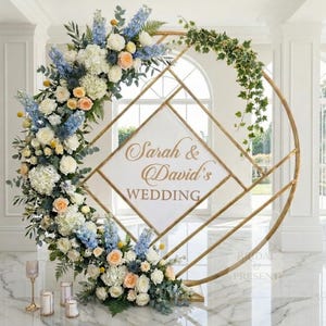 May include: A gold metal wedding arch with a geometric design, adorned with white, blue, and peach flowers and greenery. The sign in the center reads "Sarah & David's WEDDING". Candles and decorative glasses are at the base.