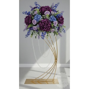 May include: A floral arrangement featuring purple hydrangeas, roses, and blue flowers. The bouquet sits atop a gold metal stand with a square base and curved supports. Strands of pearls cascade down from the flowers. The arrangement is suitable for weddings and events.