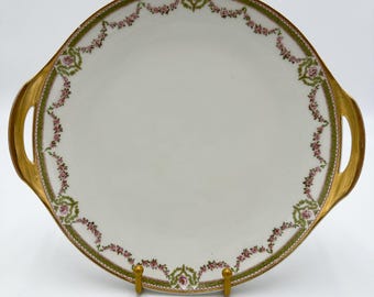 May include: A white porcelain serving platter with gold trim and a delicate floral design in pink and green. The platter has two gold handles.
