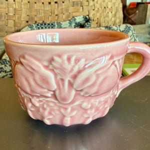 May include: A light pink ceramic mug featuring a raised, embossed relief design of two rabbits nestled among foliage and carrots. The mug has a smooth, glossy finish, a wide circular opening, and a single curved handle on the right side.