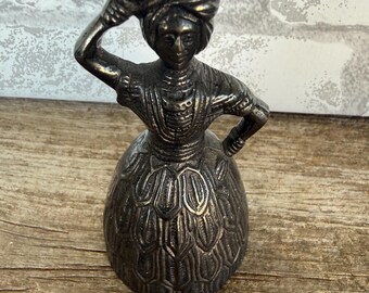 May include: A dark metallic bell shaped like a person wearing a headwrap and a textured, layered skirt. The figure has one arm raised to hold an object on their head and the other arm resting on their hip, with a detailed bodice and skirt design.