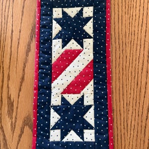 May include: A rectangular quilted table runner featuring a patriotic design with two dark blue star blocks at the top and bottom, separated by a central section of diagonal red and white stripes. The entire piece is accented with small white stars.