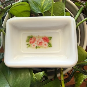 May include: A rectangular white milk glass dish featuring a central floral decal. The design displays a cluster of pink and peach-colored roses with green leaves centered on the smooth, glossy interior surface of the small, shallow container.