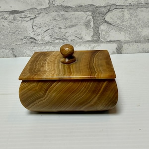 May include: A rectangular box made of polished brown and tan banded stone. The box features a smooth, curved body and a flat, removable lid topped with a small, rounded knob handle. The natural stone displays distinct, flowing horizontal layers.