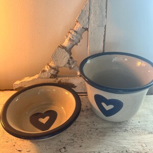 May include: Two ceramic bowls with blue rims and white interiors. Both bowls feature a blue heart with a white center. The smaller bowl is in the foreground and the larger bowl is in the background.