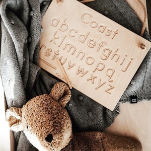 May include: A wooden alphabet tracing board with the letters of the alphabet in raised lettering. The board is light brown and has a white inscription that reads "Coast". A wooden stylus is lying on the board.