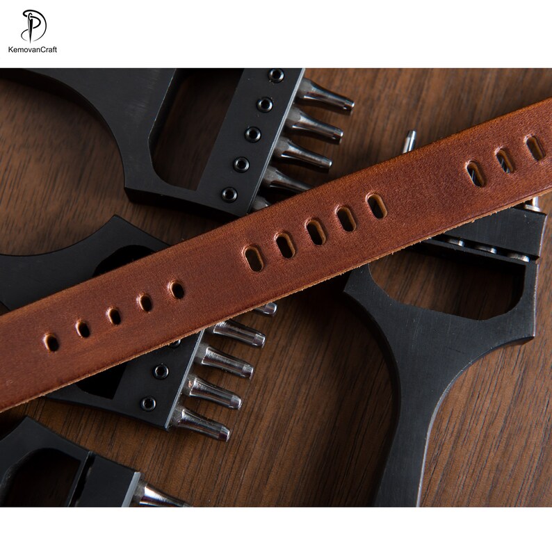 May include: Close-up of a brown leather belt with evenly spaced oval holes, lying across black metal leatherworking tools. The tools have multiple pointed prongs. The background is a wooden surface.