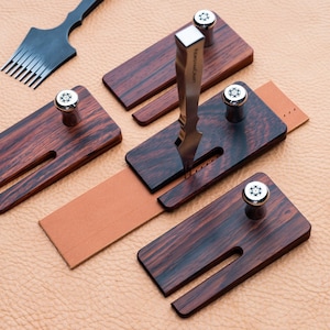 May include: Four wooden leather stitching tools with a metal awl and a stitching tool. The tools have a dark wood grain and are arranged on a light brown surface.