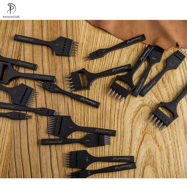 May include: A collection of black leathercraft tools, including various sizes of pronged chisels and punches. The tools are arranged on a wooden surface, showcasing their different widths and prong configurations. The brand name "KemovanCraft" is visible on each tool.