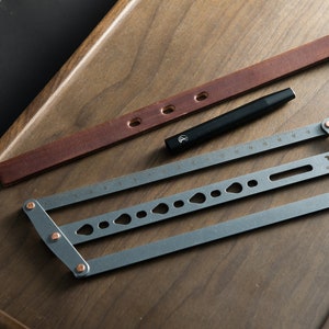May include: A gray metal tool with a ruler, holes, and a small dog silhouette. The tool is on a wooden surface with a brown leather strap and a black pen.