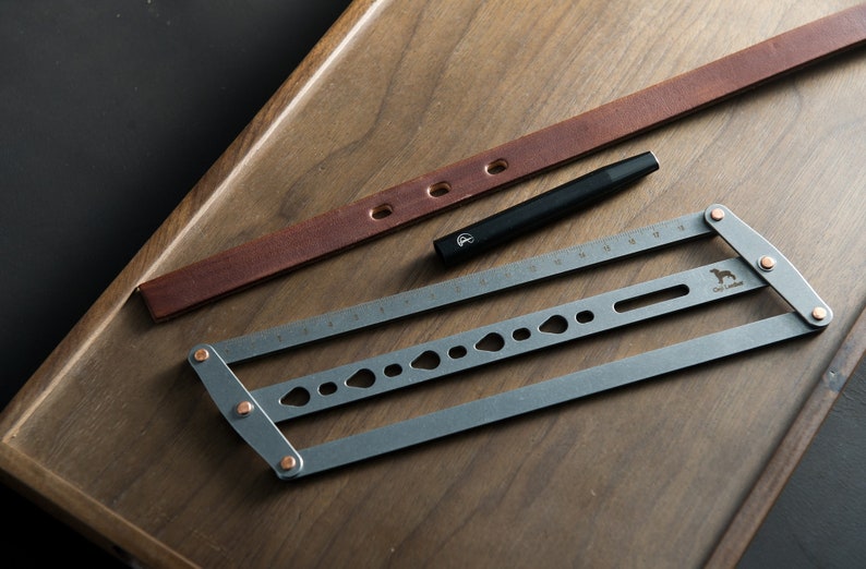 May include: A gray metal tool with a ruler, holes, and a small dog silhouette. The tool is on a wooden surface with a brown leather strap and a black pen.