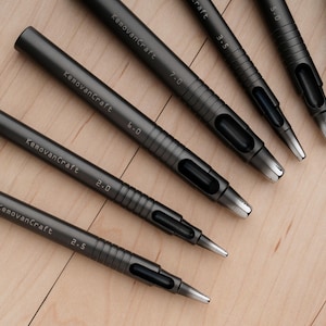 May include: A collection of dark gray leathercraft hole punches of varying sizes, arranged on a light wood surface. Each punch is labeled with the "KemovanCraft" brand name and a size measurement.