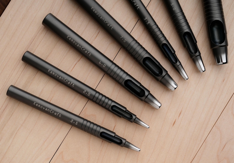 May include: A collection of dark gray leathercraft hole punches of varying sizes, arranged on a light wood surface. Each punch is labeled with the "KemovanCraft" brand name and a size measurement.