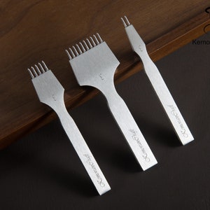 May include: Three silver leather working tools with different prong configurations. Each tool has a handle and a head with prongs for stitching leather. The tools are arranged on a dark wooden surface. The text "KemovanCraft" is printed on the handles.