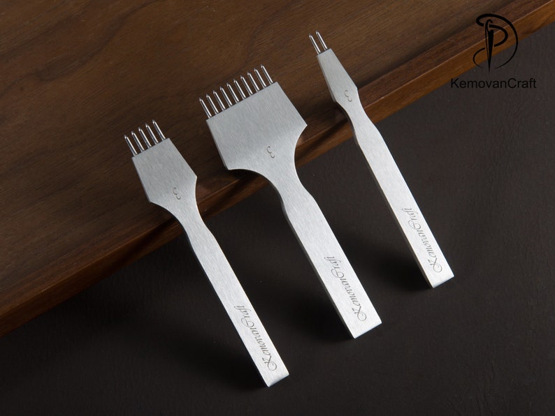 May include: Three silver leather working tools with different prong configurations. Each tool has a handle and a head with prongs for stitching leather. The tools are arranged on a dark wooden surface. The text "KemovanCraft" is printed on the handles.