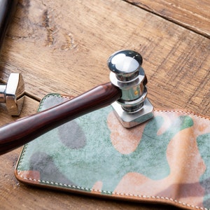 May include: A close-up of a leather working tool with a wooden handle. The tool is being used to stamp a piece of leather with a camouflage pattern.