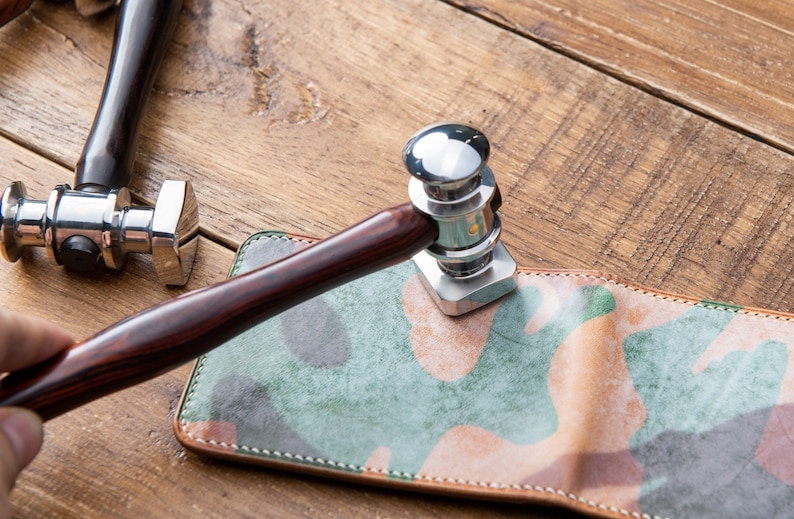 May include: A close-up of a leather working tool with a wooden handle. The tool is being used to stamp a piece of leather with a camouflage pattern.