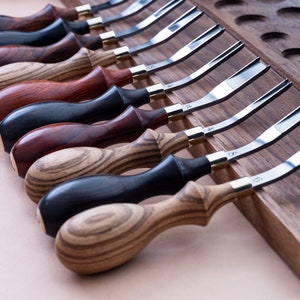 Puede incluir: Un juego de 10 herramientas de artesanía en cuero con mangos de madera. Las herramientas están dispuestas en una fila sobre una tabla de madera. Las herramientas están numeradas del 1 al 10.