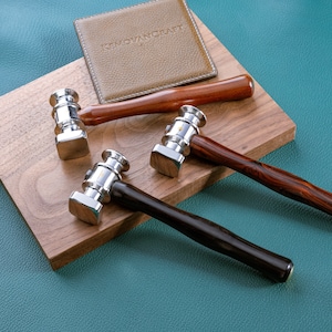 May include: Three leather working hammers with different colored wooden handles. The hammers have a silver metal head and are on a wooden surface. The text "REMOVANCRAFT" is visible on a brown leather piece in the background.