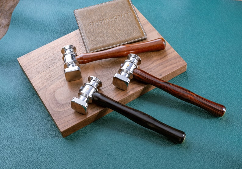 May include: Three leather working hammers with different colored wooden handles. The hammers have a silver metal head and are on a wooden surface. The text "REMOVANCRAFT" is visible on a brown leather piece in the background.