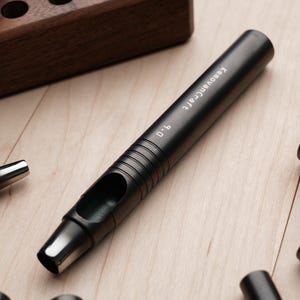 May include: A black leathercraft hole punch tool with the text "KeovanCraft 9.0" printed on the side. The tool is lying on a light wood surface, with other similar tools and a wooden storage block in the background.