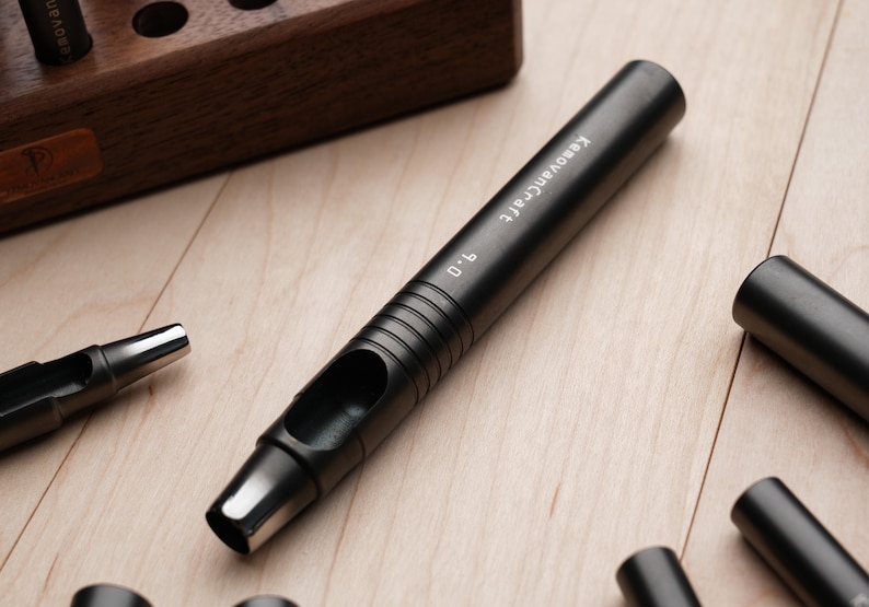 May include: A black leathercraft hole punch tool with the text "KeovanCraft 9.0" printed on the side. The tool is lying on a light wood surface, with other similar tools and a wooden storage block in the background.