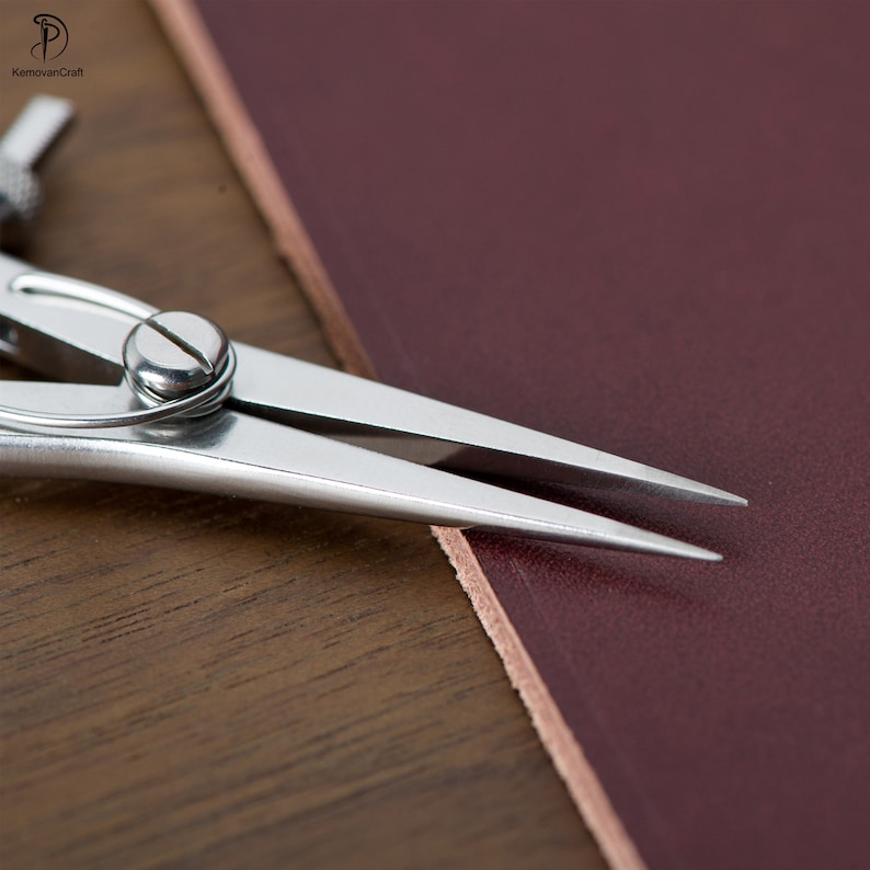 May include: A close-up of a silver leather crafting tool with a pointed tip, resting on a piece of burgundy leather. The tool is used for marking and cutting leather.