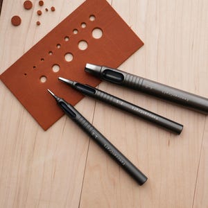 May include: Three dark gray metal leather working tools, including hole punches, are arranged on a light wood surface. A brown leather swatch with various sized holes is also present. The tools are labeled with sizes.