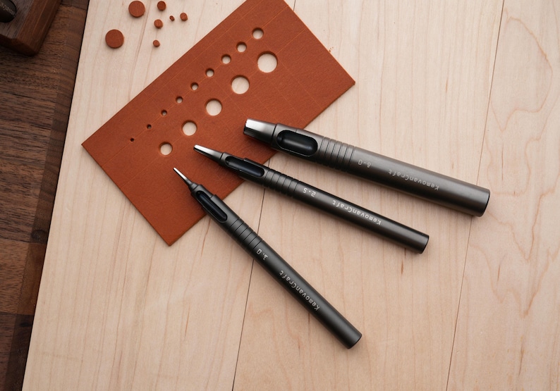 May include: Three dark gray metal leather working tools, including hole punches, are arranged on a light wood surface. A brown leather swatch with various sized holes is also present. The tools are labeled with sizes.