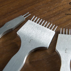 May include: Three stainless steel leather working tools with multiple prongs, each with a handle and the number "3" etched on the surface. The tools are arranged on a wooden surface. The logo "KemovanCraft" is in the upper right corner.