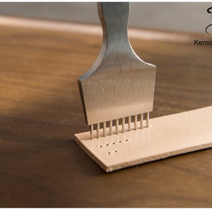 May include: A stainless steel leather punch tool with multiple prongs is pressed into a strip of light brown leather. The tool is marked with the number "3". The leather strip has a series of evenly spaced holes.