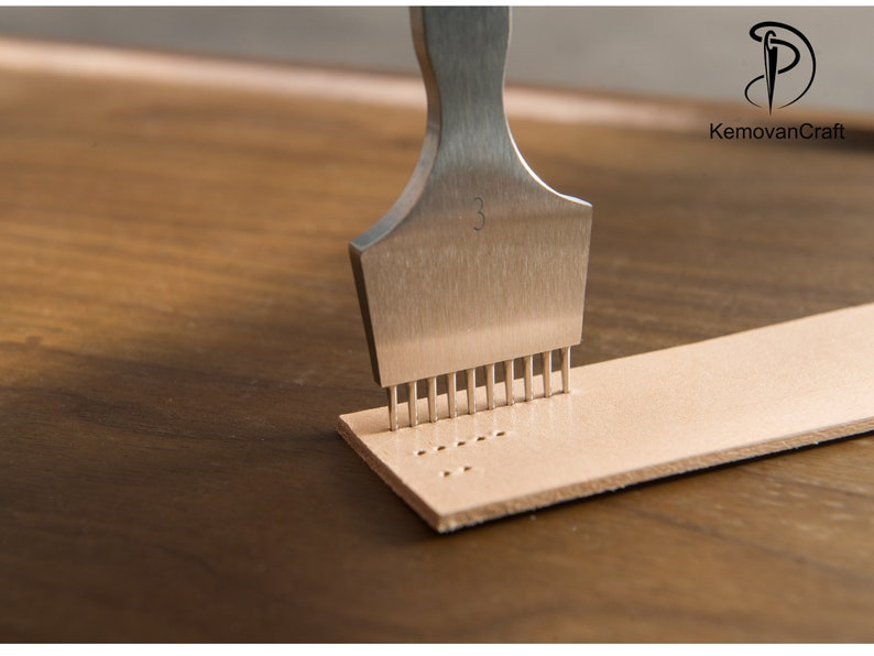 May include: A stainless steel leather punch tool with multiple prongs is pressed into a strip of light brown leather. The tool is marked with the number "3". The leather strip has a series of evenly spaced holes.