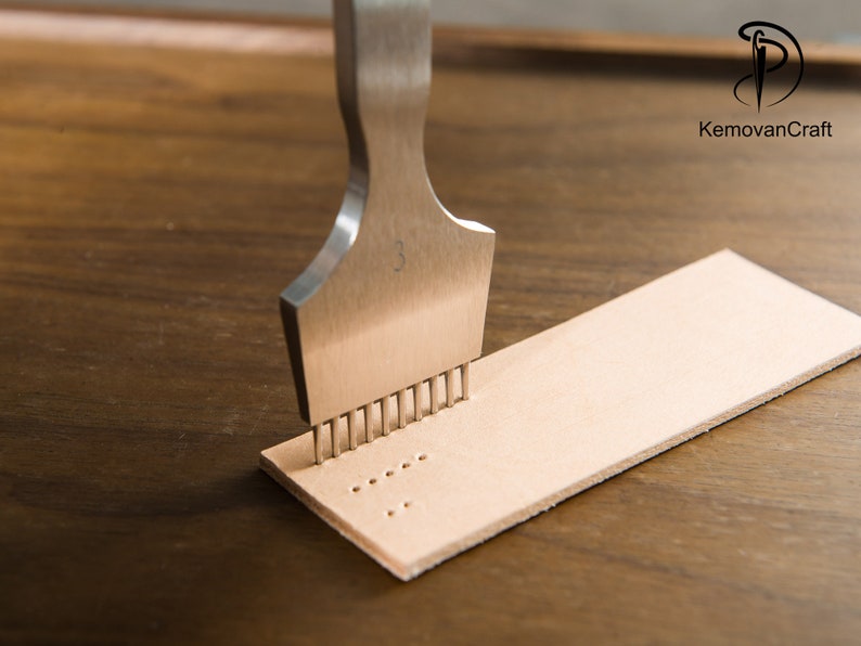 May include: A metal leathercraft tool with multiple prongs is pressed into a strip of light tan leather. The tool is marked with the number "3". The logo "KemovanCraft" is visible in the upper right corner. The leather strip is on a wooden surface.