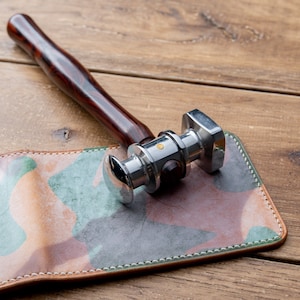 May include: A silver leather working hammer with a wooden handle resting on a camouflage patterned leather wallet.