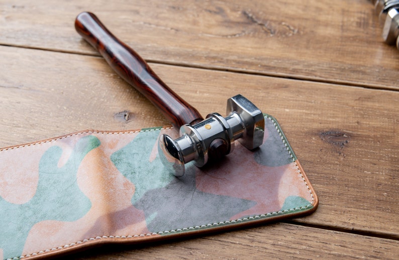 May include: A silver leather working hammer with a wooden handle resting on a camouflage patterned leather wallet.