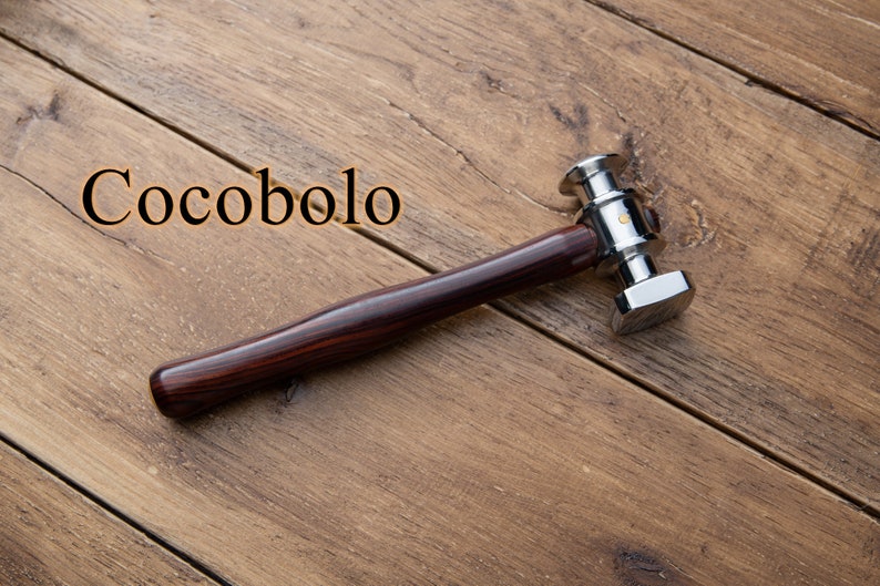 May include: A silver hammer with a wooden handle made of cocobolo wood. The hammer has a square head and a round handle. The handle is dark brown with reddish streaks. The hammer is lying on a wooden surface.