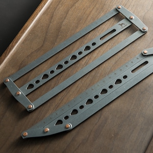 May include: Two gray metal rulers with copper accents. The rulers are folded and have various markings and holes. The rulers are on a wooden surface.