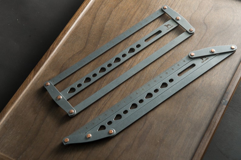 May include: Two gray metal rulers with copper accents. The rulers are folded and have various markings and holes. The rulers are on a wooden surface.