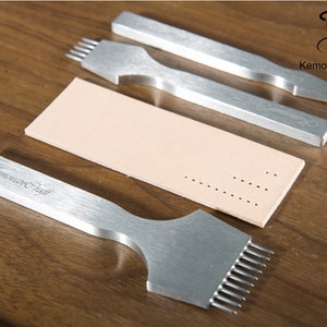 May include: A collection of silver leatherworking tools, including punches and a strip of beige leather with punched holes. The tools are arranged on a wooden surface, showcasing their design and functionality for crafting.
